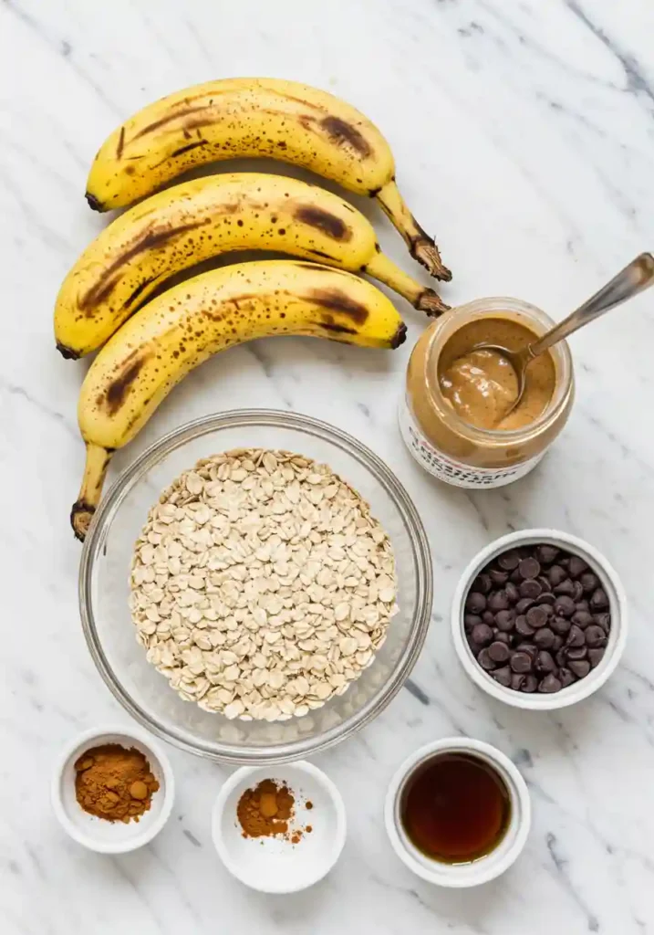 Ripe bananas, rolled oats, nut butter, maple syrup, and chocolate chips arranged on marble surface for banana oatmeal bars recipe