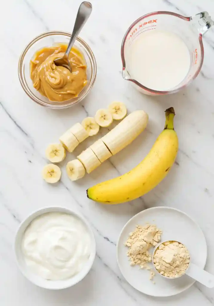 All ingredients for banana peanut butter smoothie including frozen banana chunks, natural peanut butter, Greek yogurt, almond milk, and protein powder